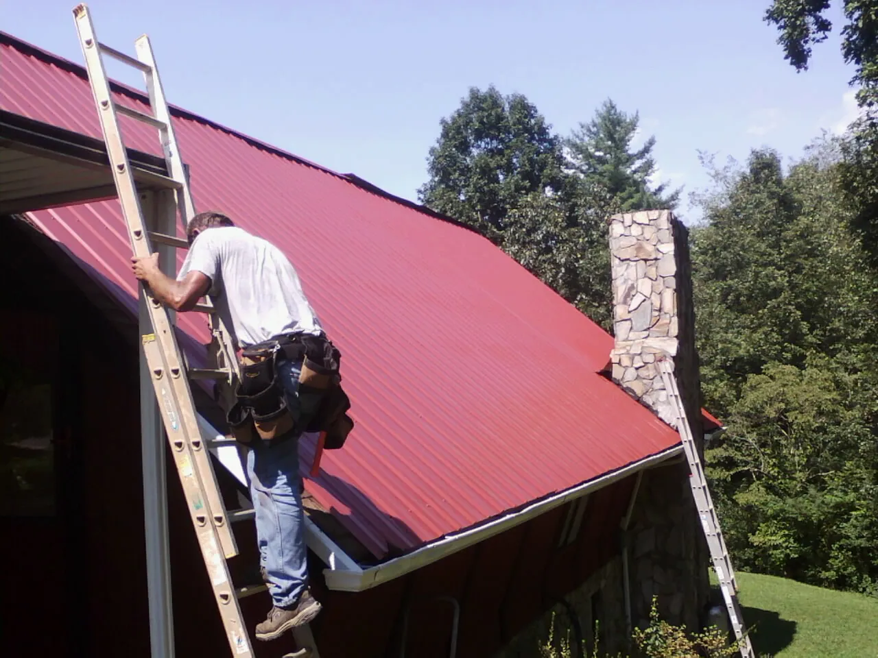 Roofer on a ladder beside a red metal roof and stone chimney — Soffit & Fascia Repair by Roofing Brothers in Oakboro